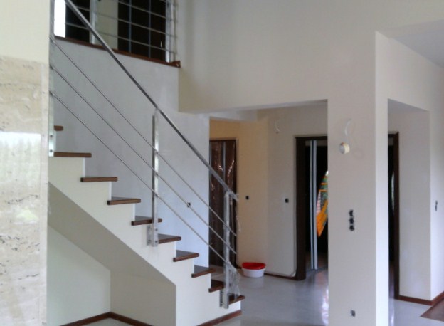 Wykończone wnętrze domu z nowoczesnymi schodami z drewnianymi stopniami i metalowymi poręczami, kremowe ściany, widoczne drzwi i fragment okna na górze.